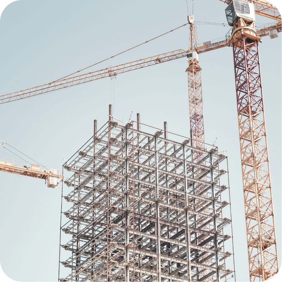 A construction site with a partially erected steel building structure, surrounded by two large cranes moving materials under a clear blue sky.