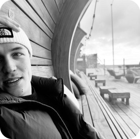 Portrait of a person in a black jacket and white beanie, sitting on a wooden bench with a relaxed expression, with a wooden wall and a terrace view in the background.