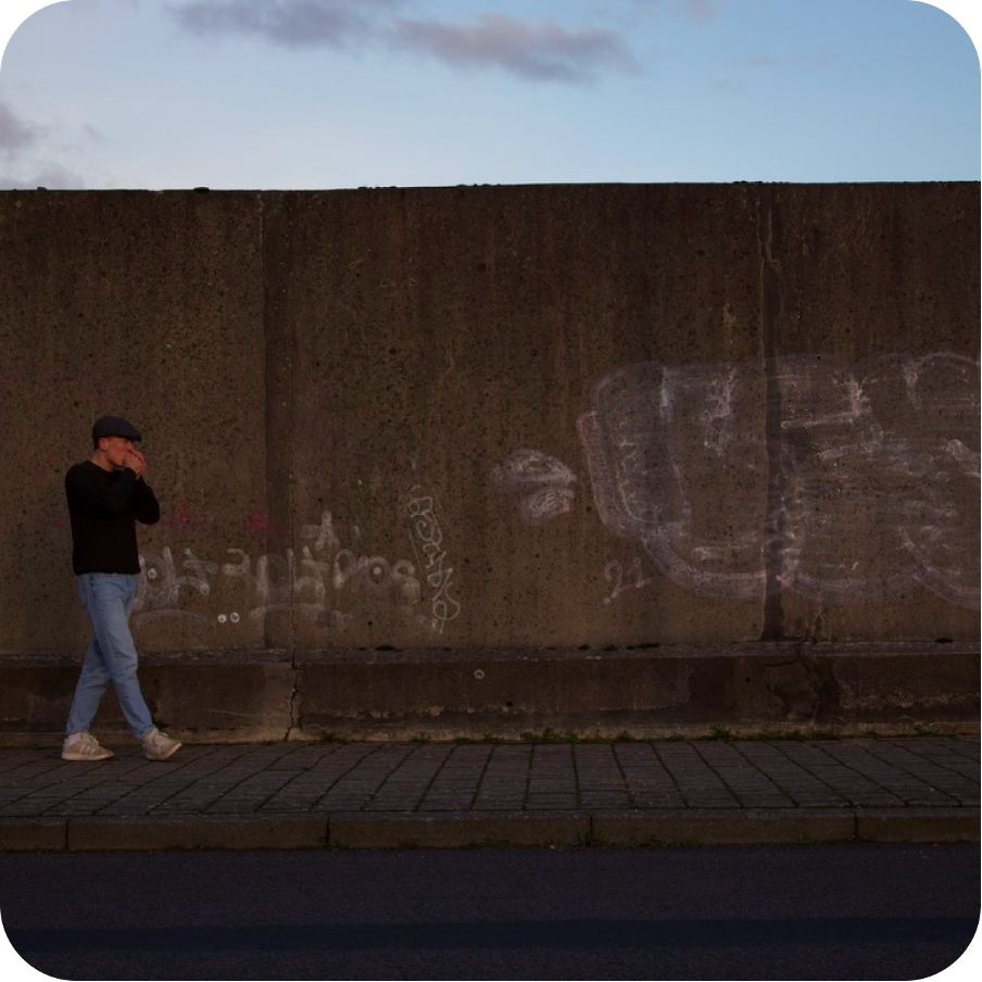 A man walking along a concrete wall with graffiti while smoking, on a paved street.